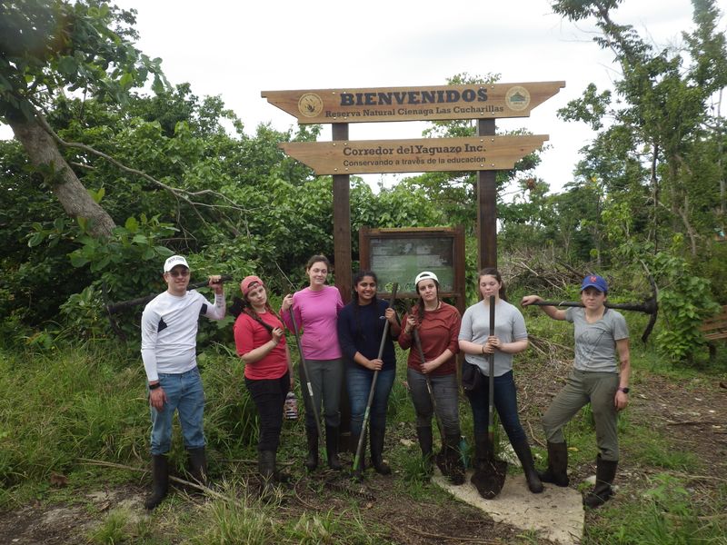 Seven students pose under a sign that says "Bienvenidos: Reserva Natural Cienaga Las Cucharillas"