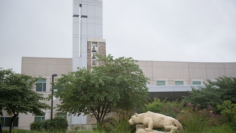 A statue of the Penn State Nittany Lion at the Penn State Health St. Joseph campus appears in profile in front of a row of trees and weeds. Behind the scene rises a glass tower and bells and windows.