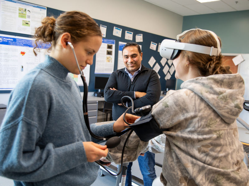 Student takes blood pressure of another student on stationary bike wearing VR goggles while faculty observes