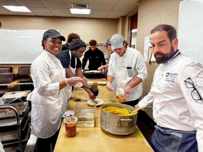 Six students and an instructor wearing chef coats spoon polenta onto plates