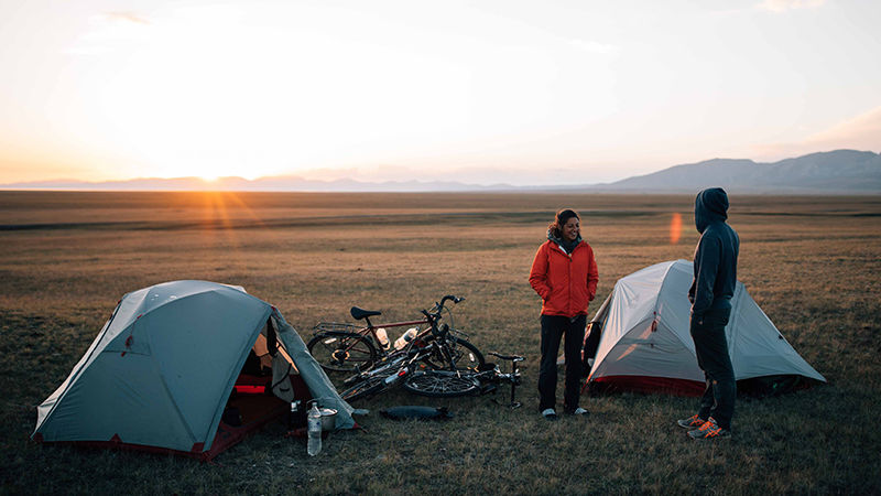 Nicole and a companion stand among their small white tents and a heap of bicycles on a grassy field, with the sun rising in the distance