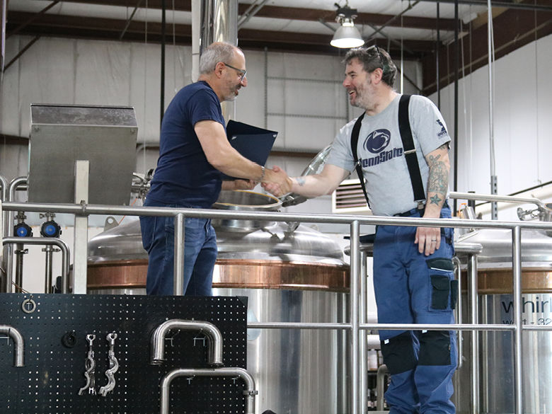 An instructor and a student shake hands in front of some brew kettles