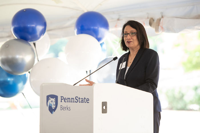 Penn State President Neeli Bendapudi speaks at a podium displaying the Penn State Berks mark, with blue and white balloons in the background. 