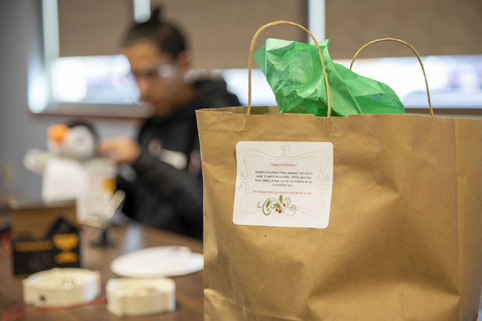 A gift bag with green tissue paper sits on a table in the foreground with a high school student assembling a button in the background.