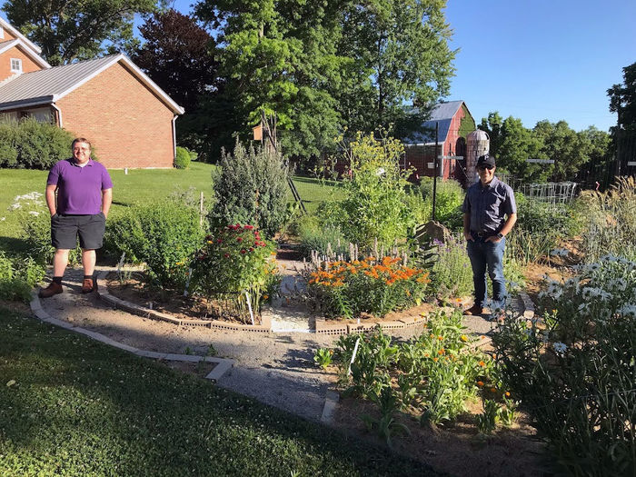 Two men stand beside a collection of plants in the sun