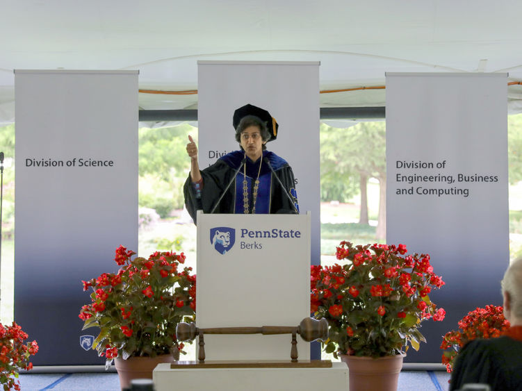 Radha Pyati, wearing robes and a cap, speaks at a podium outside under a tent at a Convocation